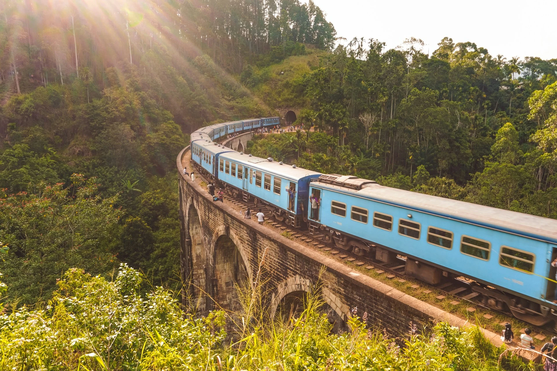 A blue passenger train traveling across the curved stone Nine Arch Bridge surrounded by lush green hills and sunbeams.