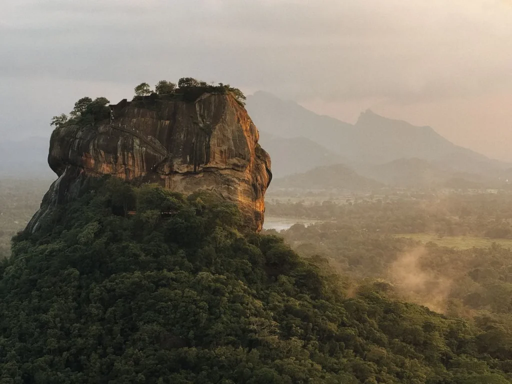 The ancient Sigiriya rock fortress rising above a dense tropical forest with misty mountains in the background during golden hour.