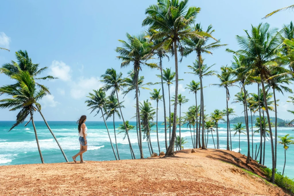 A woman walking along the reddish-brown crest of Coconut Tree Hill surrounded by tall palm trees overlooking turquoise ocean waves.