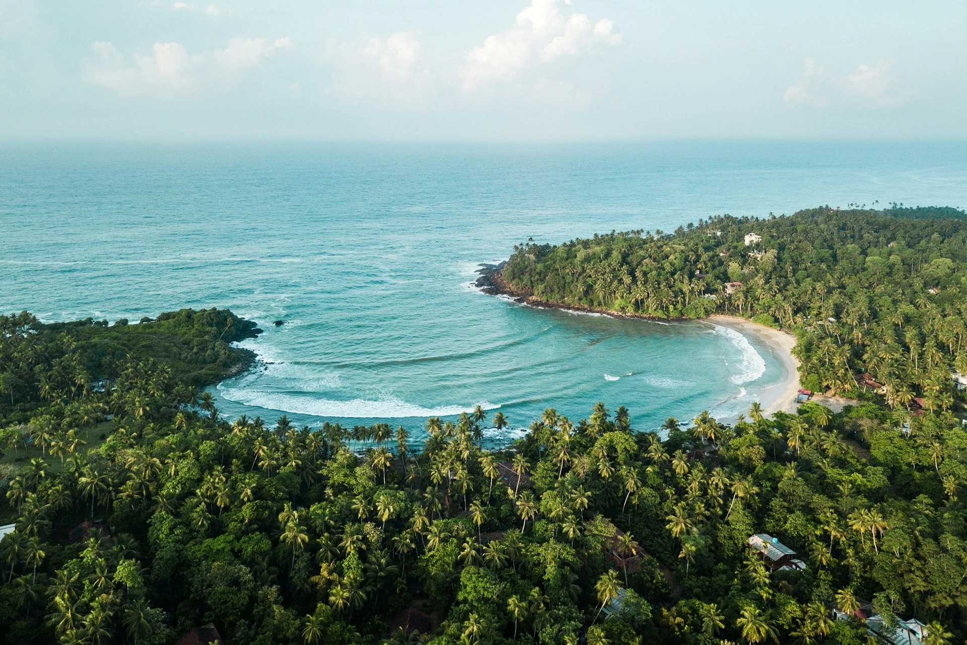 Aerial view of a crescent-shaped tropical bay with gentle ocean waves, bordered by a sandy beach and dense green palm tree forest.