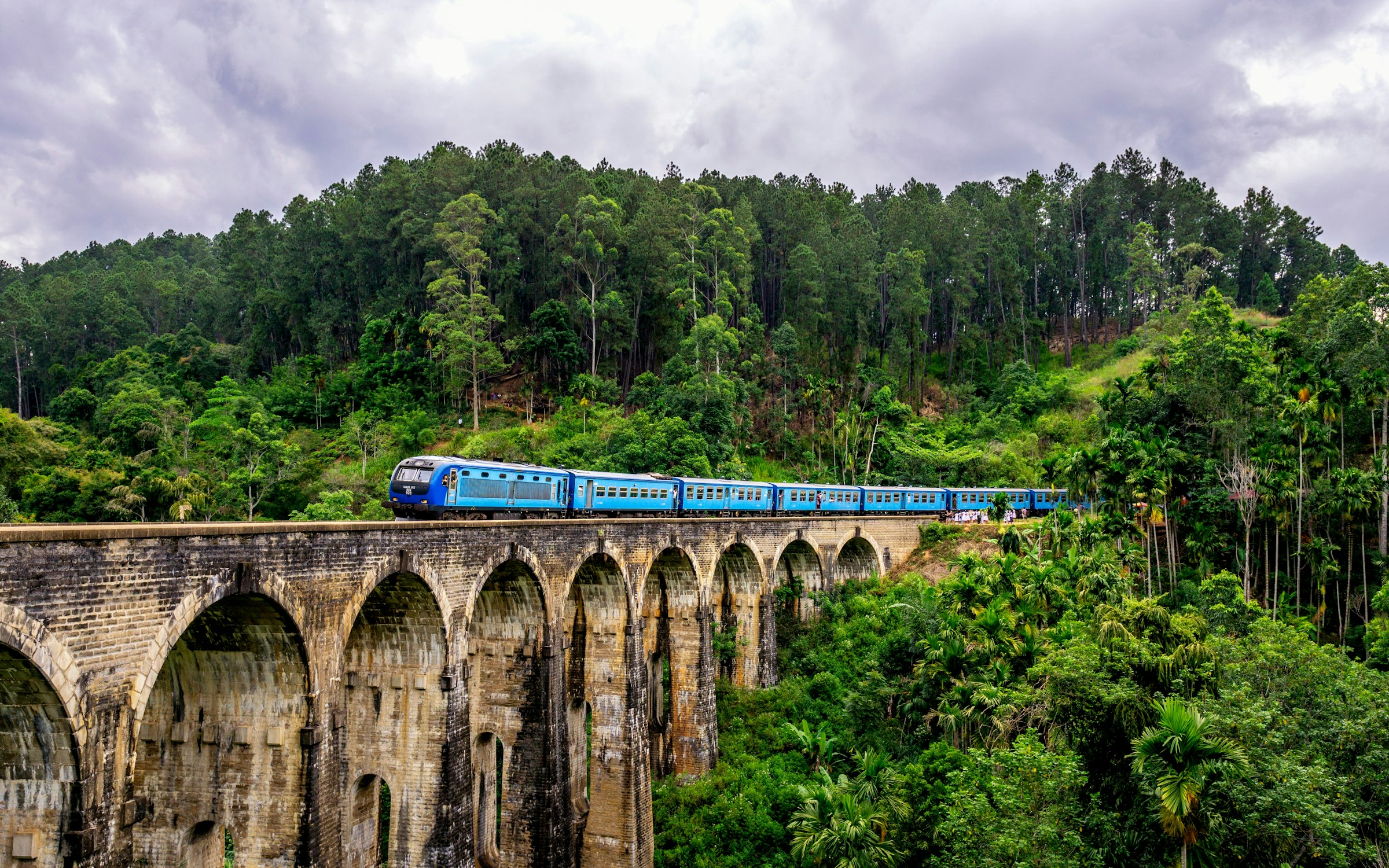 Blue train crossing the historic Nine Arch Bridge surrounded by lush green hills in Ella, Sri Lanka